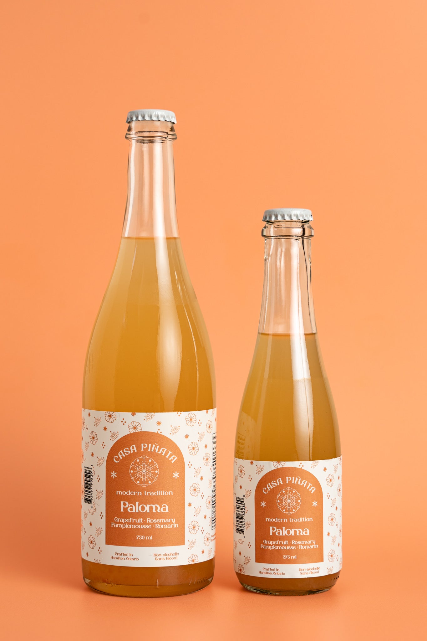 Two bottles of Casa Piñata Paloma Mocktail, one 375ml and one 750ml clear champagne bottle, against a pink background.