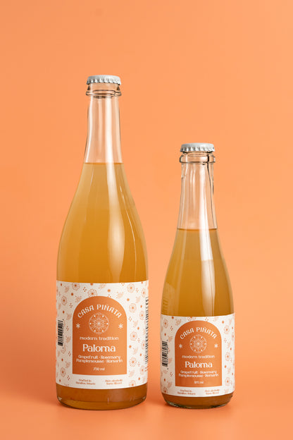 Two bottles of Casa Piñata Paloma Mocktail, one 375ml and one 750ml clear champagne bottle, against a pink background.