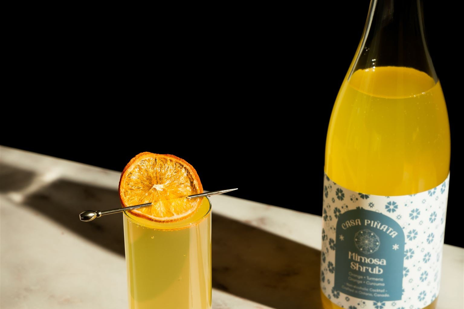 Cocktail with an orange slice in a tall glass next to a bottle of yellow liquid on a dark surface.